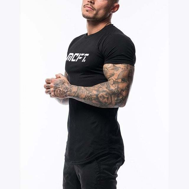 Muscular man wearing black short sleeve MCFT Shirts with white logo and tattooed arms against white background